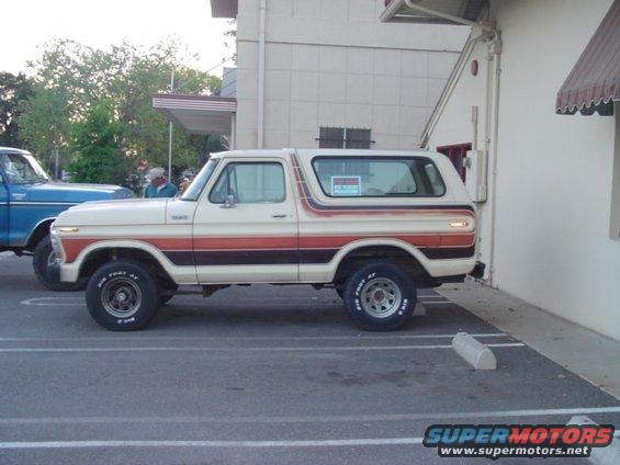 Side view of a cream-colored Ford Bronco with orange and brown stripes. A blue Ford truck is parked next to it