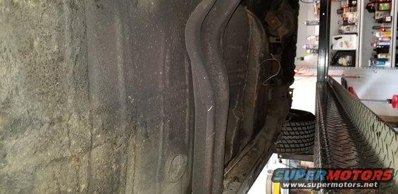 Underside of a vehicle showing fuel tank and exhaust system, with tire and lift.