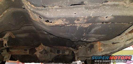 Underside of a car showing severe rust and holes in the frame and body.