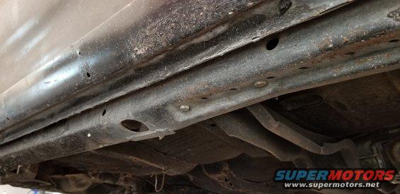 Underside of a vehicle showing rusted frame rails and suspension components.