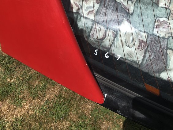 Red spoiler attached to the rear of a black car, obscuring part of the window with a patterned curtain and handwritten numbers 5, 6, 7