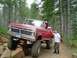 1976 Ford F-100 - A man poses with a lifted red Ford pickup truck navigating a rocky off-road path in a forest.