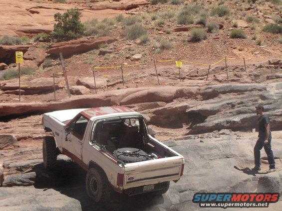White Ford Bronco with red trim driving over rocks, spare tire in the back. Person walks nearby