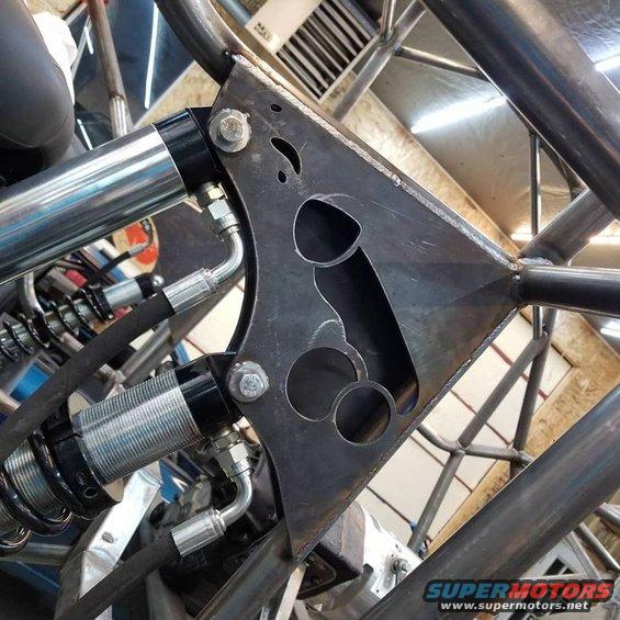 Custom metal bracket with a penis-shaped cutout, attached to shock absorbers and roll cage tubing