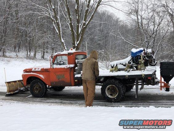 Orange 1961 Ford F-350 truck with a snow plow and a spreader on the back, parked in the snow
