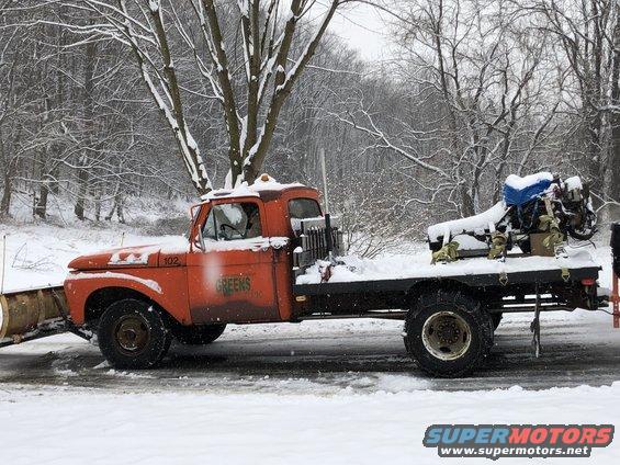 Orange truck with a snow plow attachment and a flatbed carrying an engine, in a snowy environment