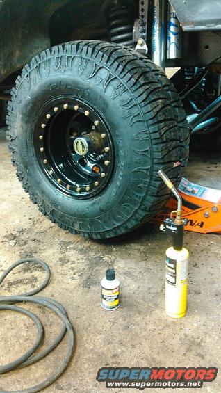 Off-road tire on black beadlock wheel with gold bolts, jack, and torch
