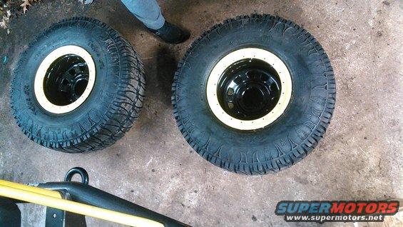 Two off-road tires with black rims and cream trim rings on concrete