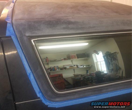 Dark car roof and window frame covered in blue masking tape, reflecting a workshop interior