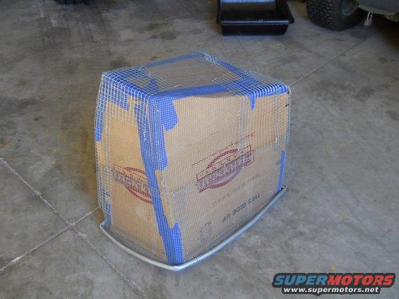 Cardboard box covered in wire mesh and blue tape, with a metal base, on a concrete floor