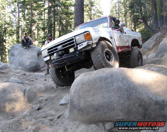 White and red Ford Bronco off-roading over rocks, front grille visible