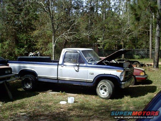 Silver and blue Ford F-150 pickup truck with hood open, parked in a grassy area with trees