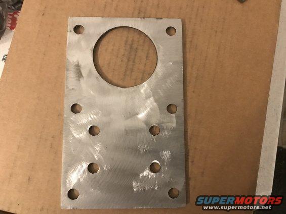 Rectangular metal plate with a large central hole and eight smaller surrounding holes, on a brown surface