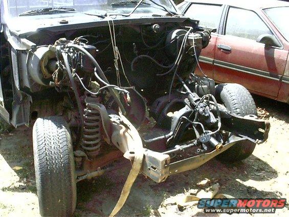 Front chassis and engine bay of a disassembled vehicle, with a red sedan in the background