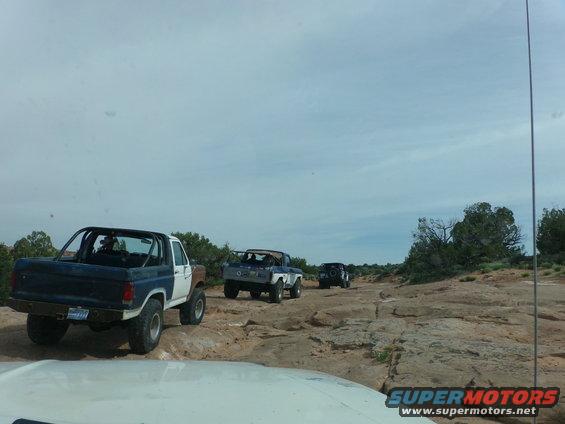 Several off-road vehicles, including a blue and white Bronco, drive on a rocky trail under a cloudy sky