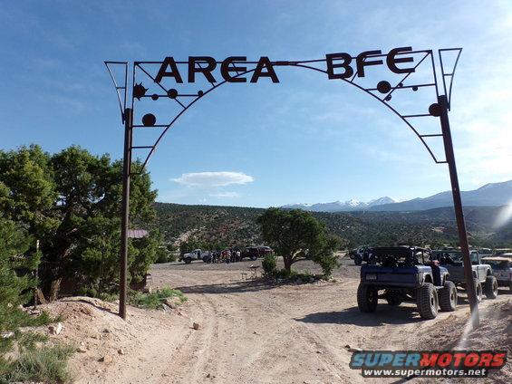 Blue Ford Bronco with large tires parked on dirt road, "AREA BFE" arch overhead