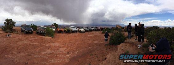 Group of off-road vehicles parked on a rocky, dusty area with people standing nearby; storm clouds gather in the distance