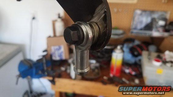 Close-up of a metal rod end joint with a bolt and washer, likely part of a vehicle's steering or suspension system. Background shows a workshop with tools and supplies