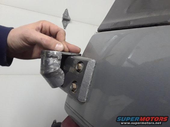Hand holding a metal bracket attached to a gray surface, possibly a vehicle part