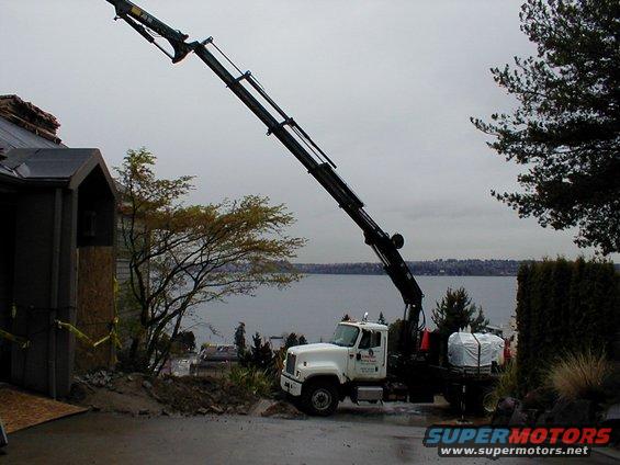 White crane truck with boom extended, parked on wet pavement near a house under construction. Water and trees in background
