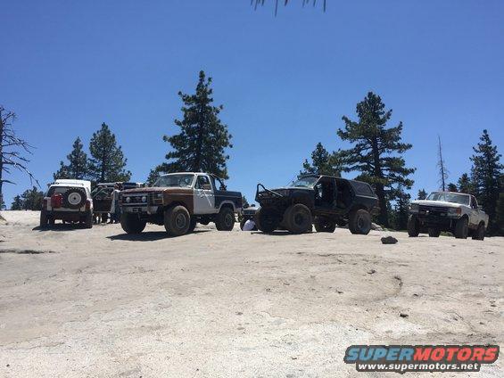 Several modified off-road trucks and SUVs parked on a dusty, rocky surface under a clear blue sky