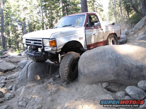 White and maroon Ford F-Series truck with a winch, navigating large rocks on a trail