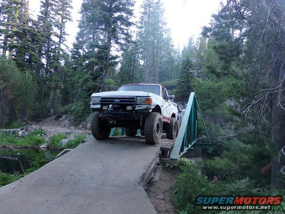 White pickup truck with off-road tires crossing a bridge. Body style suggests 1980s Ford F-Series