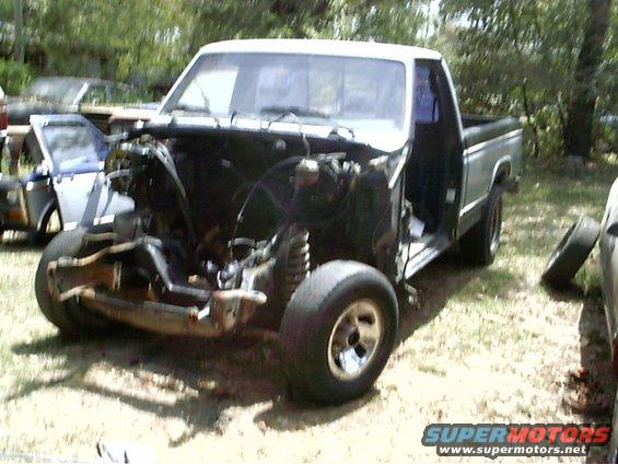 Front end of a pickup truck with engine removed, showing suspension and chassis. White cab, silver body trim