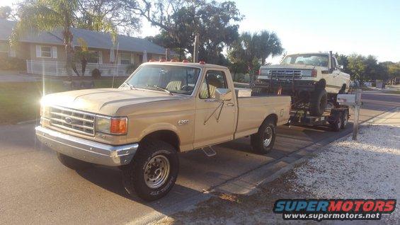 Tan Ford F-Series pickup truck, towing a white lifted truck on a trailer