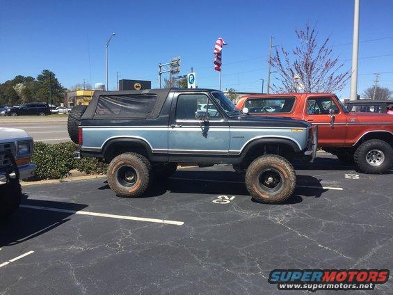 Side view of a lifted blue Ford Bronco with a soft top, parked next to an orange Ford Bronco