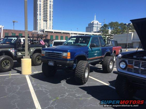 Lifted blue and teal Ford pickup truck with large tires. License plate reads 475VMR. Other modified vehicles and buildings in background