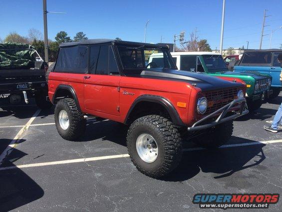 Red Ford Bronco with black hood and soft top, lifted suspension, aftermarket bumper. Other vehicles visible behind it