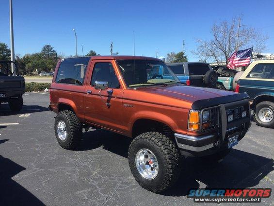 Orange Ford Bronco II with aftermarket wheels and tires, parked in a lot with other vehicles