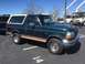 1979 Ford Bronco - A green Ford Bronco with a tan stripe and a white removable top is parked in a lot with utility trucks in the background.