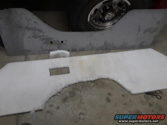 Gray vehicle interior panel with cutouts and white insulation material, next to a silver wheel