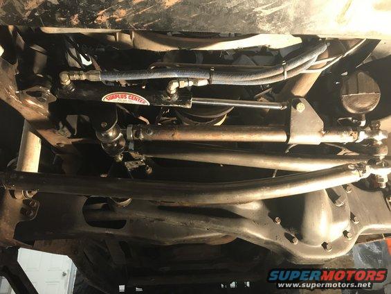 Underside view of vehicle steering linkage and front axle with a 'Surplus Center' sticker on a hydraulic cylinder