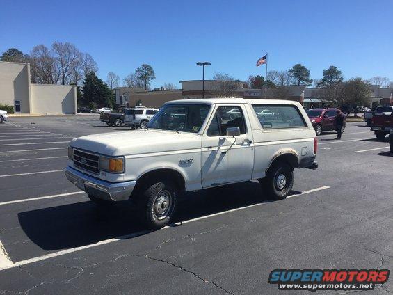 White Ford Bronco parked in a lot, side view. Other vehicles and buildings in background