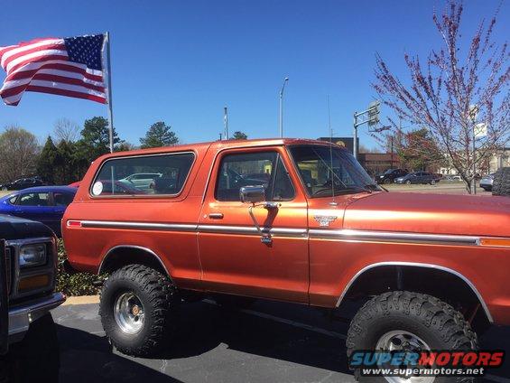 Orange Ford Bronco with large tires parked outside, American flag flying nearby