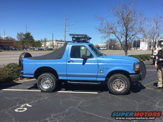 Blue Ford Bronco with a black soft top, roof rack, and spare tire on the back. The vehicle has a bull bar on the front