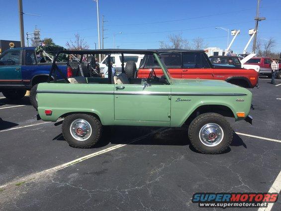 Green 1979 Ford Bronco parked in a lot, side view. Red Ford Bronco and blue truck in background
