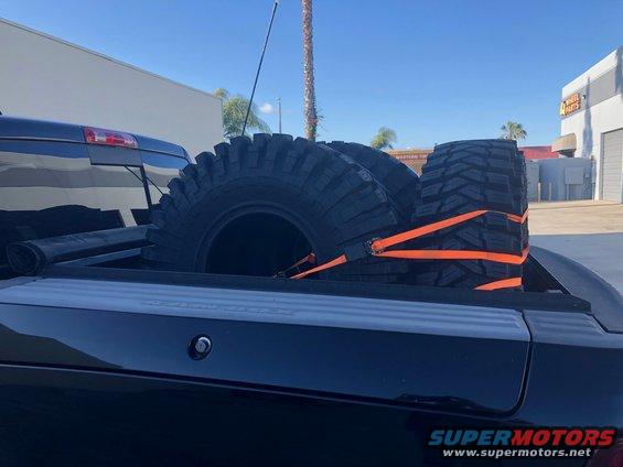 Three large off-road tires secured with orange straps in the bed of a black pickup truck