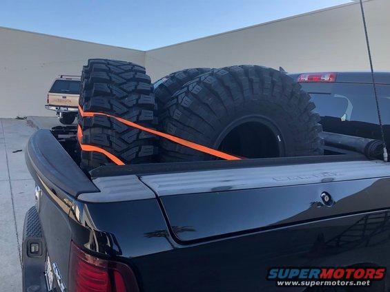 Four large off-road tires secured with orange straps in the bed of a black pickup truck. Another pickup truck is visible in the background
