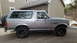 1995 Ford Bronco - This silver Ford Bronco is shown from the side, parked in front of a house with a spare tire on the back.