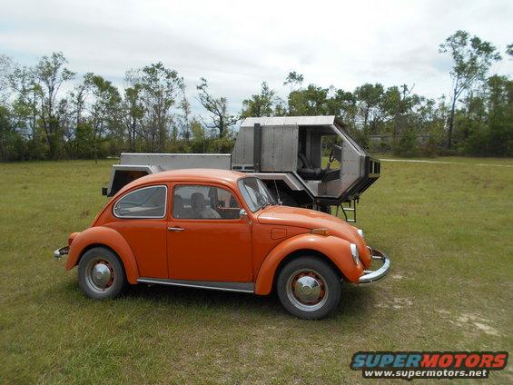 Orange Volkswagen Beetle parked on grass next to a metallic trailer with a futuristic cabin