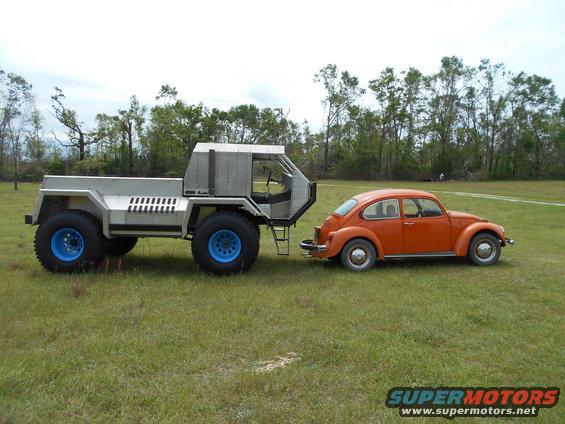 Custom-built truck with large blue wheels towing an orange Volkswagen Beetle on a grassy field
