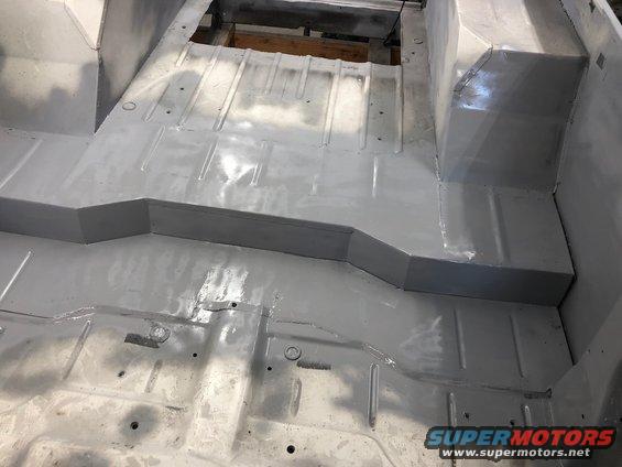 Gray painted vehicle floor with visible ribs and mounting holes, showing the transmission tunnel and seat mounts
