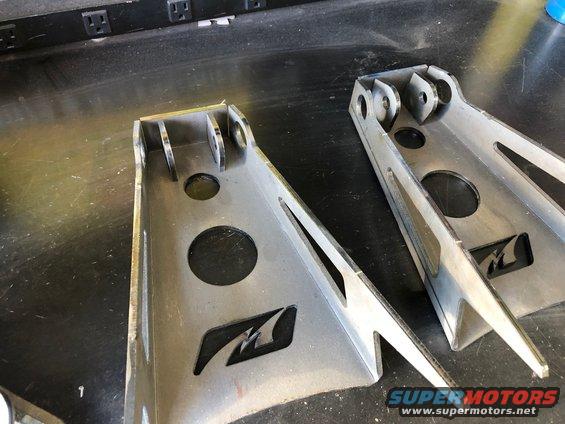 Two metal suspension brackets with a cutout logo on a workbench