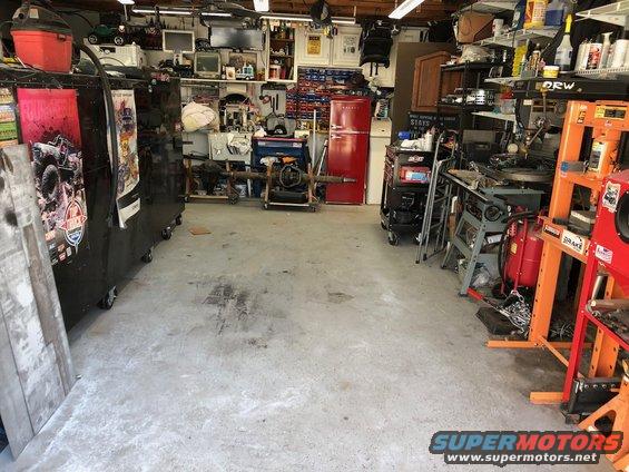Garage interior with tool chests, a red refrigerator, and various equipment. A drivetrain is on a stand in the center