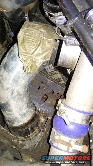 Engine bay showing a large metal pipe, a blue silicone coupler, and a yellow plastic bag covering some components. A label is visible on a metal bracket