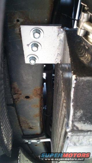 Custom aluminum bracket with three bolts attached to a rusty frame rail, next to a radiator
